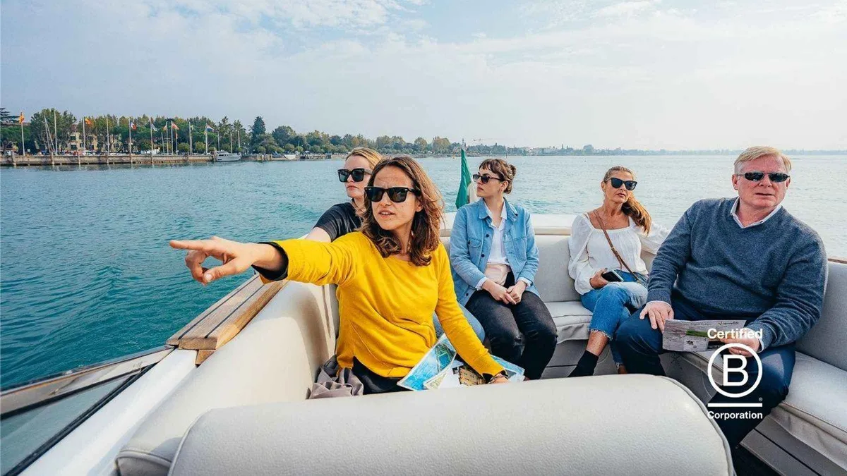 Four travellers on the white cushioned deck of a motorboat, one pointing toward the Sirmione shoreline