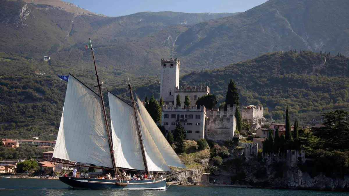 A two-masted wooden sailboat with white sails passing below Malcesine castle and its tower